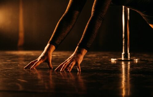 Dancer's hands on a lit stage floor during pole dancing floor work transitions, chrome pole visible in background